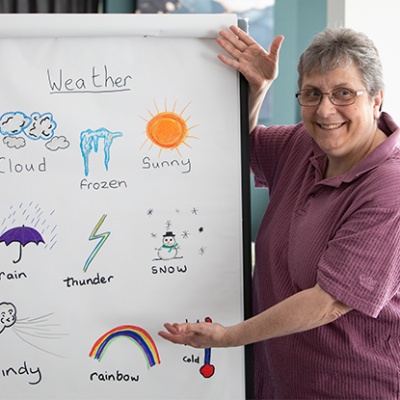 A photograph of customer Linda teaching a sign language class about the weather for residents of one of our retirement living communities. Linda has drawn symbols for weather conditions on a flip chart, and is gesturing towards the symbols with a smile.